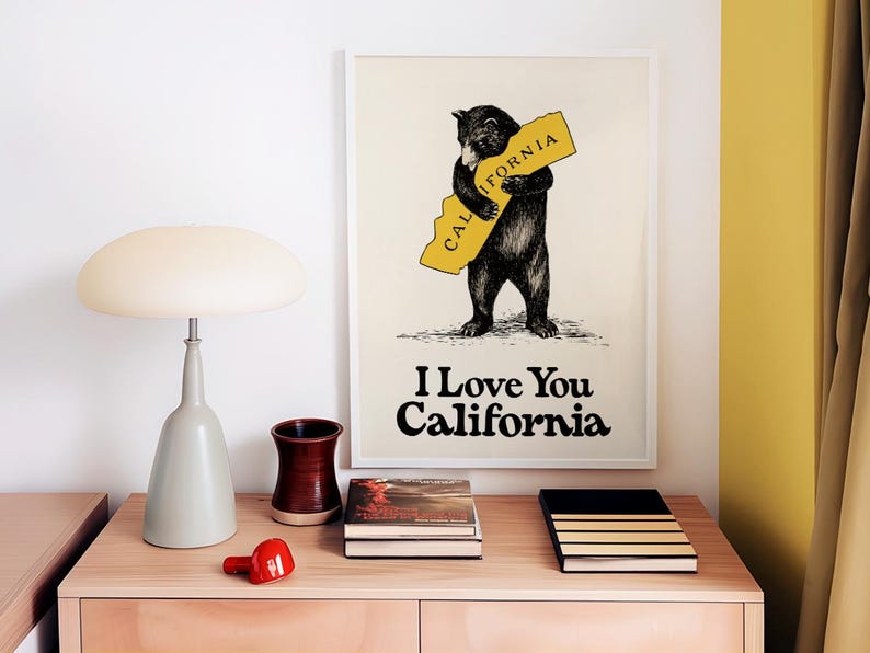 May include: A framed print with a black bear hugging a yellow outline of the state of California. The text "I Love You California" is printed below the bear. The print is on a wooden table with books and a lamp.