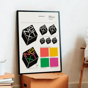 May include: A framed poster with a black border features the word "NEXT" in a 3D cube design. The design is repeated in various sizes and colors, with a color palette below. A white mug and books are in the foreground.