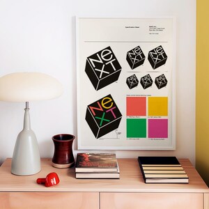 May include: A framed poster with the word "NEXT" in a cube design, along with color swatches, sits above a wooden cabinet. A gray lamp, a red object, a brown vase, and books are on the cabinet.