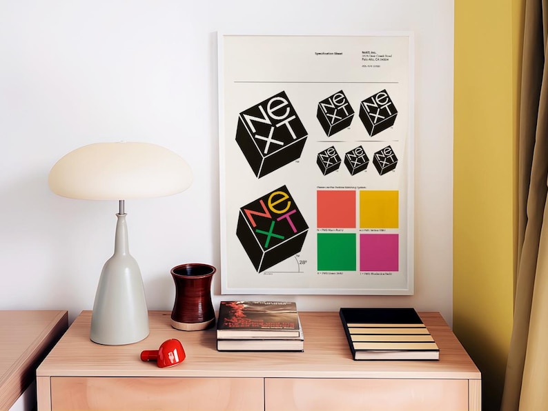 May include: A framed poster with the word "NEXT" in a cube design, along with color swatches, sits above a wooden cabinet. A gray lamp, a red object, a brown vase, and books are on the cabinet.