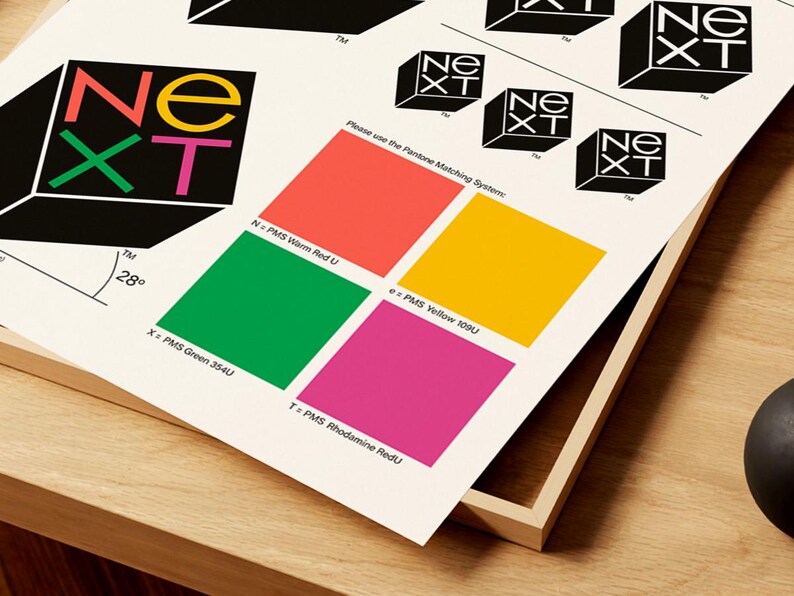 May include: A print featuring the "NEXT" logo in a 3D cube design, with color swatches and text. The logo uses red, green, yellow, and pink. The text includes "Please use the Pantone Matching System:" and color names.