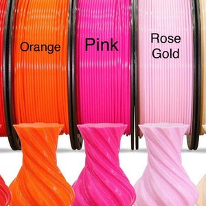 May include: Spools of orange, pink, and rose gold 3D printing filament are displayed above matching twisted vase-shaped objects. The spools are labeled with their respective colors. The vases have a spiral design.