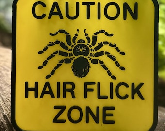 May include: Yellow warning sign with black text and a black silhouette of a spider. The sign reads "CAUTION HAIR FLICK ZONE".