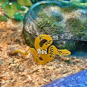 May include: A gold-colored, snake-shaped decorative ornament featuring intricate cut-out leaf patterns along its body. The word Koi is written in white, cursive script across the center of the snake figure, which is positioned horizontally.