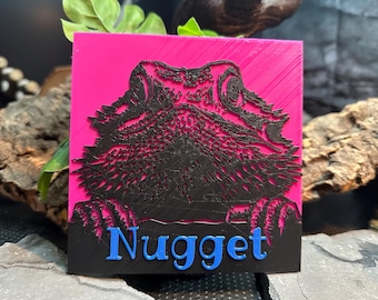 May include: A pink square tile with a black silhouette of a bearded dragon's head. The dragon's eyes are visible and the tile has the word "Nugget" written in blue letters at the bottom.