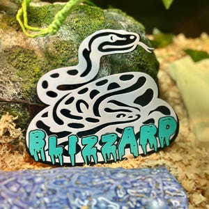 May include: A white and black snake-shaped pin with the word "BLIZZARD" in teal dripping letters. The pin is set against a backdrop of a mossy rock and wood shavings.