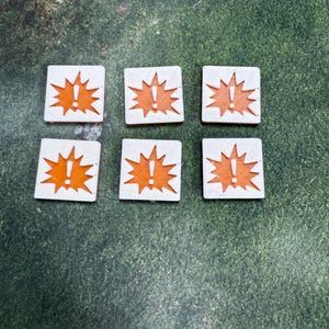 May include: Six square game tokens with a white background and an orange exclamation point inside an explosion shape. The tokens are arranged on a green surface.