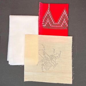 May include: An assortment of fabric pieces, including a folded white cloth, a red square with embroidered green and white designs, and a beige square with a floral embroidery pattern. The edges of the red and beige squares are unfinished.