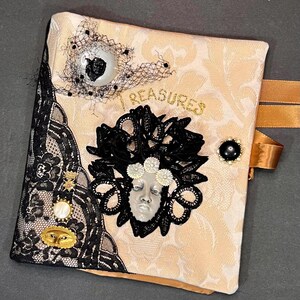 May include: A decorative journal with a cream-colored damask pattern, embellished with black lace, a black and white eye-shaped button, and a face-shaped cameo. The word "TREASURES" is written in gold script. A gold ribbon is attached.