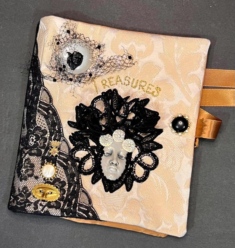 May include: A decorative journal with a cream-colored damask pattern, embellished with black lace, a black and white eye-shaped button, and a face-shaped cameo. The word "TREASURES" is written in gold script. A gold ribbon is attached.