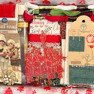 May include: A Christmas-themed craft kit in a box, filled with red and green embroidery floss, ribbon, and vintage ephemera. Includes a card with children, a red paper cutout with carolers, and a tag from "The Faerywood Tree".