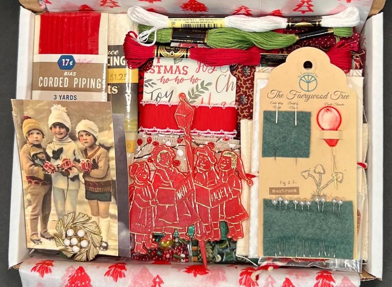 May include: A Christmas-themed craft kit in a box, filled with red and green embroidery floss, ribbon, and vintage ephemera. Includes a card with children, a red paper cutout with carolers, and a tag from "The Faerywood Tree".
