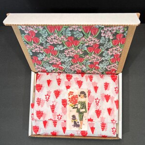 May include: An open cardboard box with a floral patterned interior. Inside, tissue paper with a red and white design covers a product. A small sticker with a vintage illustration of a woman and a dog is visible.
