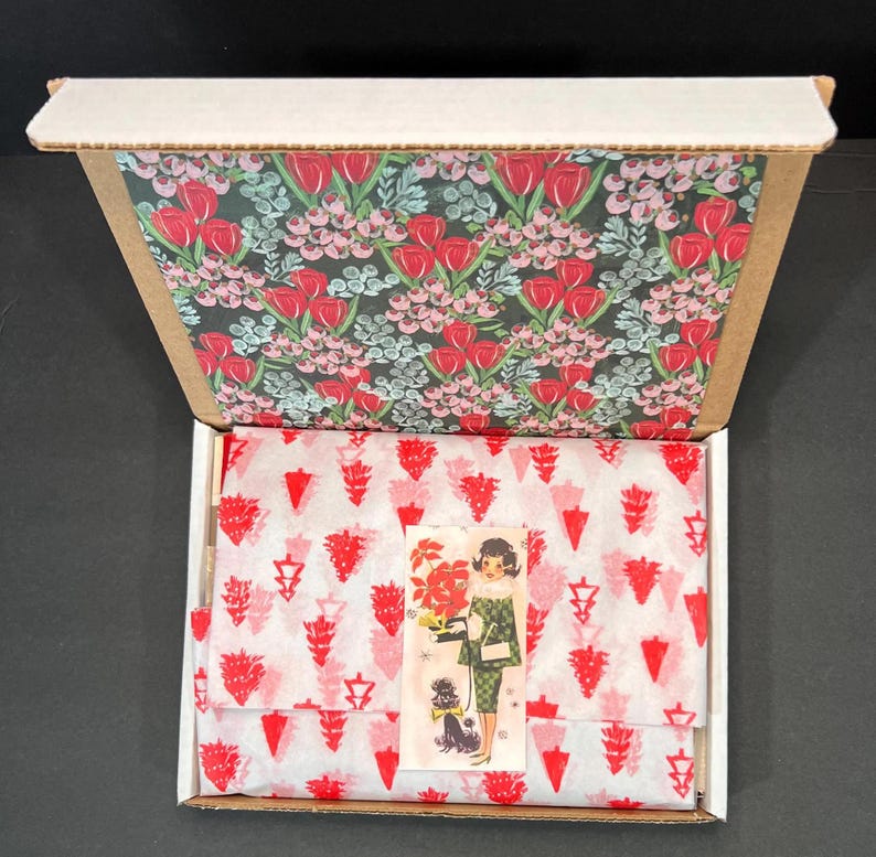 May include: An open cardboard box with a floral patterned interior. Inside, tissue paper with a red and white design covers a product. A small sticker with a vintage illustration of a woman and a dog is visible.