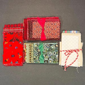 May include: A collection of fabric bundles in various patterns and colors. Red, green, and cream fabrics are tied with ribbons and twine. The fabrics include floral, paisley, and geometric designs, suitable for quilting or crafting projects.