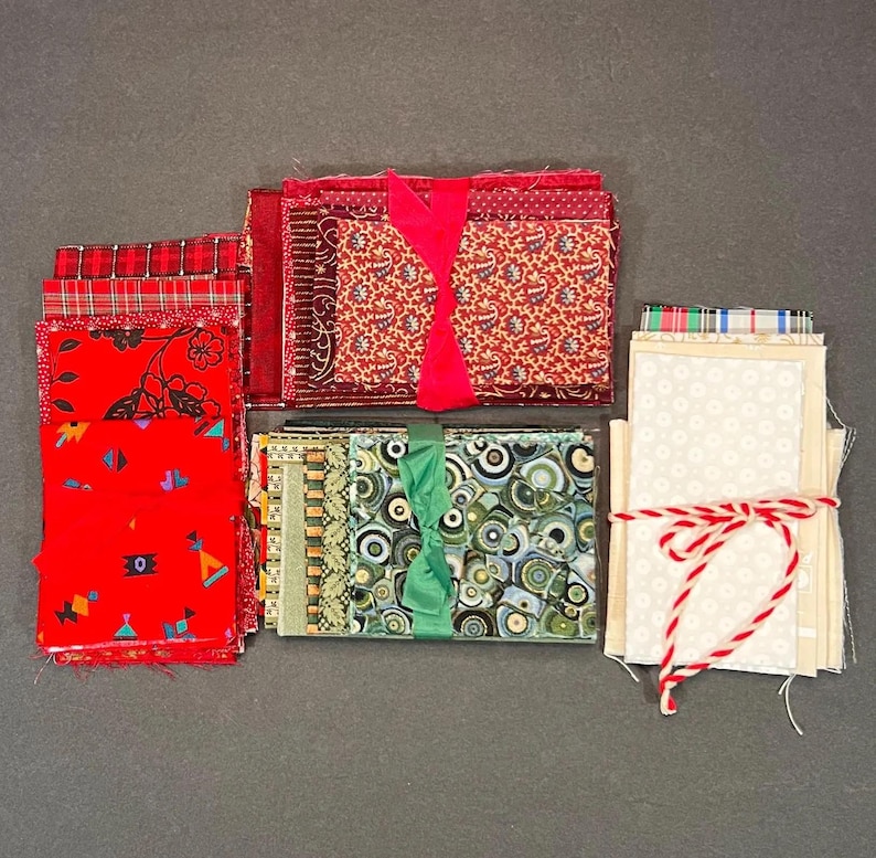 May include: A collection of fabric bundles in various patterns and colors. Red, green, and cream fabrics are tied with ribbons and twine. The fabrics include floral, paisley, and geometric designs, suitable for quilting or crafting projects.