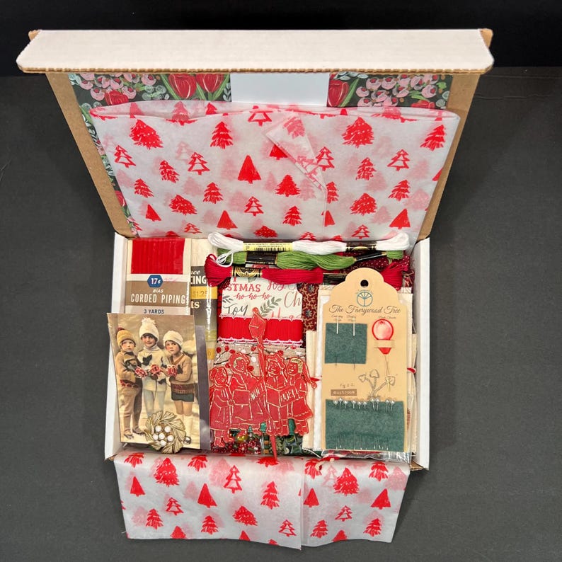 May include: An open cardboard box filled with Christmas-themed craft supplies. The box contains red and white tissue paper with Christmas tree patterns, embroidery floss, corded piping, and a vintage Christmas card.