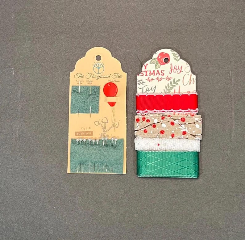 May include: Two decorative tags. One features needles and the text "The Faerywood Tree". The other tag has a stack of Christmas ribbons in red, white, and green. The top ribbon has the word "Joy".