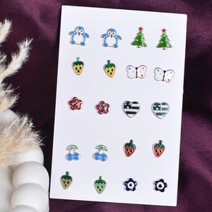 Puede incluir: Una colección de pendientes de botón coloridos sobre una tarjeta blanca. Los pendientes presentan varios diseños, incluyendo pingüinos, árboles de Navidad, fresas, mariposas, flores, corazones, cerezas y formas florales. Los pendientes son de varios colores, incluyendo azul, verde, rojo y amarillo.