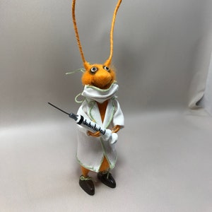 May include: A felt orange creature with long orange antennae wearing a white lab coat and a face mask, holding a syringe.