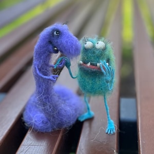 Pode incluir: Dois brinquedos de monstros de feltro, um azul e outro verde, estão sentados em uma superfície de madeira. O monstro azul tem um pescoço longo e uma cabeça pequena com um único olho. O monstro verde tem um corpo redondo com dois olhos grandes e uma boca larga.