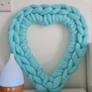 Puede incluir: Una corona de coraz&oacute;n de punto de color azul claro colgada en una pared blanca. Un difusor de aceites esenciales de madera y blanco est&aacute; sentado en una superficie blanca en primer plano.