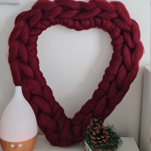 Puede incluir: Una gran corona de coraz&oacute;n tejida de color burdeos que cuelga de una pared blanca. Un peque&ntilde;o arreglo de pi&ntilde;as en maceta y un difusor blanco con base de madera est&aacute;n sobre una superficie blanca debajo de la corona.