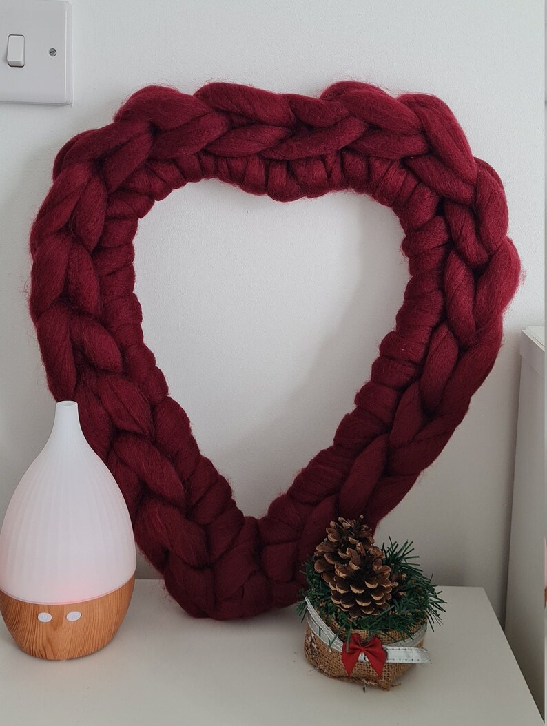 Puede incluir: Una gran corona de coraz&oacute;n tejida de color burdeos que cuelga de una pared blanca. Un peque&ntilde;o arreglo de pi&ntilde;as en maceta y un difusor blanco con base de madera est&aacute;n sobre una superficie blanca debajo de la corona.