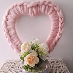 Puede incluir: Una corona de coraz&oacute;n de lana rosa con un ramo de flores blancas y melocot&oacute;n en un jarr&oacute;n plateado.