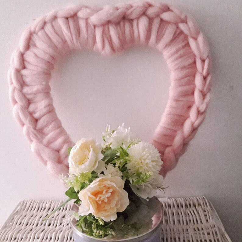 Puede incluir: Una corona de coraz&oacute;n de lana rosa con un ramo de flores blancas y melocot&oacute;n en un jarr&oacute;n plateado.