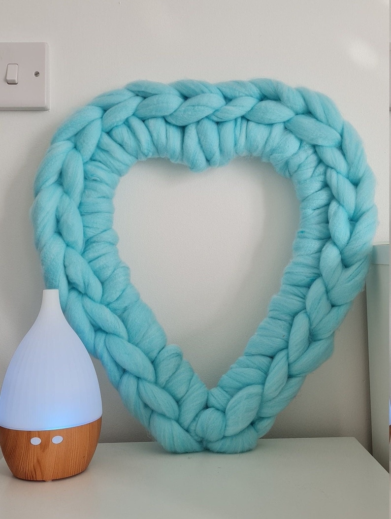 Puede incluir: Una corona de coraz&oacute;n de punto de color azul claro colgada en una pared blanca. Un difusor de aceites esenciales de madera y blanco est&aacute; sentado en una superficie blanca en primer plano.