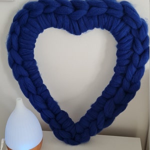 Puede incluir: Una gran corona de coraz&oacute;n tejida en azul, hecha con hilo grueso, cuelga de una pared blanca.