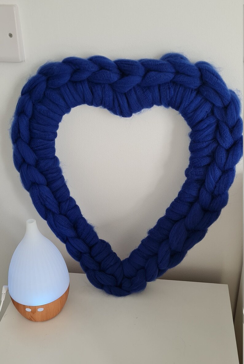 Puede incluir: Una gran corona de coraz&oacute;n tejida en azul, hecha con hilo grueso, cuelga de una pared blanca.