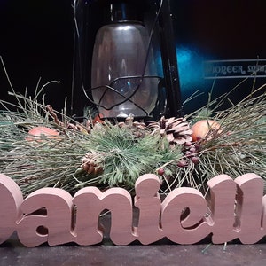 May include: A wooden sign with the name "Danielle" spelled out in large, pink letters. The sign is sitting on a table with a green wreath and a lantern in the background.