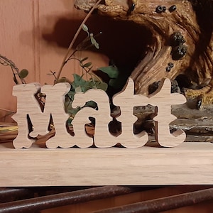 May include: Wooden name sign with the name "Matt" carved in a decorative font. The sign is on a wooden base and is sitting on a rustic surface.