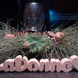 May include: A wooden sign with the name "LaVonne" carved in cursive letters. The sign is painted a light brown color and has a heart-shaped cutout at the end. The sign is sitting on a dark surface.