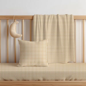 May include: A square throw pillow and a rectangular blanket featuring a tan and white gingham check pattern. A crescent moon-shaped plush object hangs from the wooden crib railing. The bedding set displays a consistent checkered design.