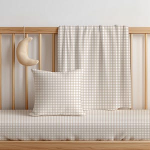 Könnte beinhalten: Ein quadratisches Kissen und eine rechteckige Decke mit beigefarbenem und weißem Vichy-Karo-Muster. Ein halbmondförmiges Plüschtier hängt am hölzernen Gitterbett. Das Bettwäsche-Set zeigt durchgehend ein einheitliches Karomuster.