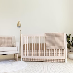 Puede incluir: Una cuna blanca con una falda de cuna de vichy marrón y blanco y una manta a juego. Una mecedora blanca con una almohada de vichy marrón y blanco se encuentra delante de la cuna. Una planta en maceta se encuentra en el suelo junto a la cuna.
