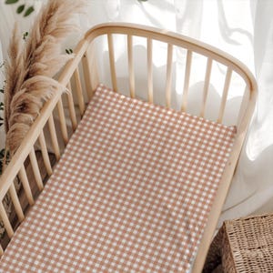 May include: A cot sheet featuring a repeating terracotta and white gingham check pattern. The fabric is fitted over a rectangular mattress inside a light-toned wooden cot with vertical slats, creating a clean and uniform textile appearance.