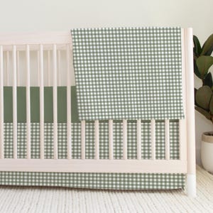 May include: A light-colored wooden crib featuring a sage green and white gingham patterned blanket draped over the side, a solid sage green crib sheet, and a matching sage green and white gingham patterned crib skirt covering the base.