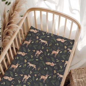 May include: A dark gray crib sheet featuring a repeating pattern of brown deer, fawns, green ferns, and small mushrooms. The fabric is stretched over a light-colored wooden crib mattress frame, displaying a woodland-themed illustration.