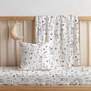 May include: A white square throw pillow and a matching white blanket featuring a repeating watercolor pattern of gray rabbits, green stems, and small blue berries. A crescent moon-shaped plush object hangs from the wooden crib railing.