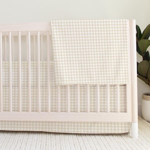 Puede incluir: Una cuna de madera de color claro con barrotes verticales y un protector de tela con estampado de cuadros vichy en beige y blanco. Una manta a juego con el mismo estampado de cuadros vichy beige y blanco cuelga del borde superior.