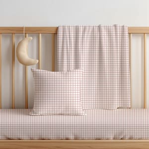 May include: A light pink and white gingham patterned square cushion, a matching rectangular blanket, and a fitted sheet, all arranged on a wooden cot frame. A small, crescent moon-shaped plush toy hangs from the side of the cot railing.