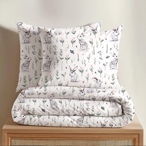 Può includere: Un set di biancheria da letto bianco con un motivo ripetuto di conigli grigi, steli verdi e piccoli fiori rosa. Il set comprende un piumino piegato e due federe abbinate, tutte decorate con la stessa stampa botanica e animale.