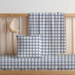 Könnte beinhalten: Ein quadratisches Kissen, eine rechteckige Decke und ein Spannbettlaken, alle mit einem blau-weißen Vichy-Karo-Muster. Ein kleines, halbmondförmiges Plüschobjekt hängt am hölzernen Gitterbettgeländer in der Nähe des Kissens.