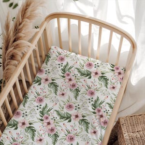 May include: A crib sheet featuring a repeating pattern of dusty pink roses, green fern leaves, and small white floral sprigs on a white background, fitted over a light-toned wooden crib frame with vertical slats and a curved headboard.