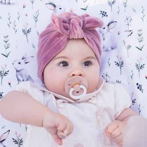 Peut inclure: Un turban pour b&eacute;b&eacute; en velours rose poudr&eacute; avec un n&oelig;ud sur le devant, accompagn&eacute; d'une t&eacute;tine rose clair. Le bonnet est port&eacute; sur une chemise blanche &agrave; col volant&eacute;, sur un fond en tissu blanc orn&eacute; d'un motif r&eacute;p&eacute;titif de lapins et de fleurs.