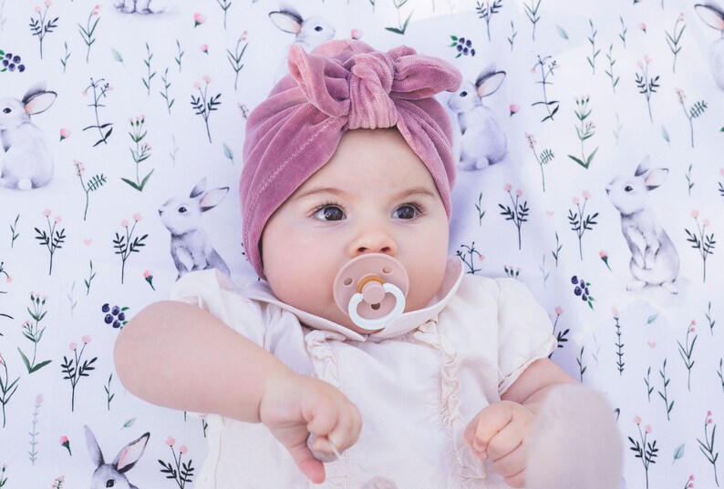 Peut inclure: Un turban pour b&eacute;b&eacute; en velours rose poudr&eacute; avec un n&oelig;ud sur le devant, accompagn&eacute; d'une t&eacute;tine rose clair. Le bonnet est port&eacute; sur une chemise blanche &agrave; col volant&eacute;, sur un fond en tissu blanc orn&eacute; d'un motif r&eacute;p&eacute;titif de lapins et de fleurs.